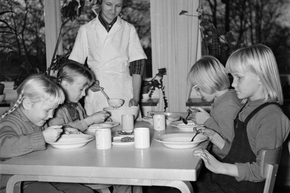 Koululaiset ruokapöydän äärellä ruokailemassa keittäjän katsoessa vieressä.